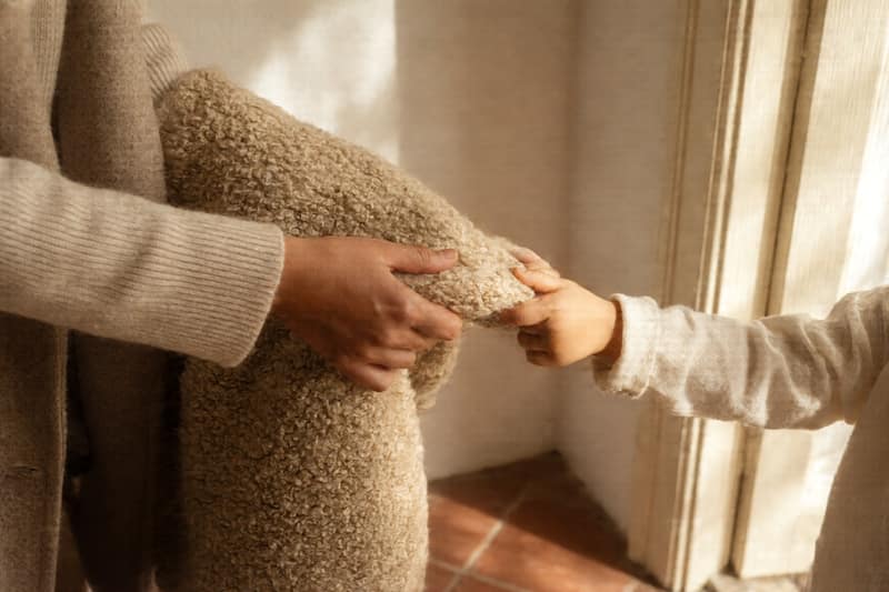 Deux mains, une adulte et une petite, tiennent ensemble un manteau près d’une porte ensoleillée.